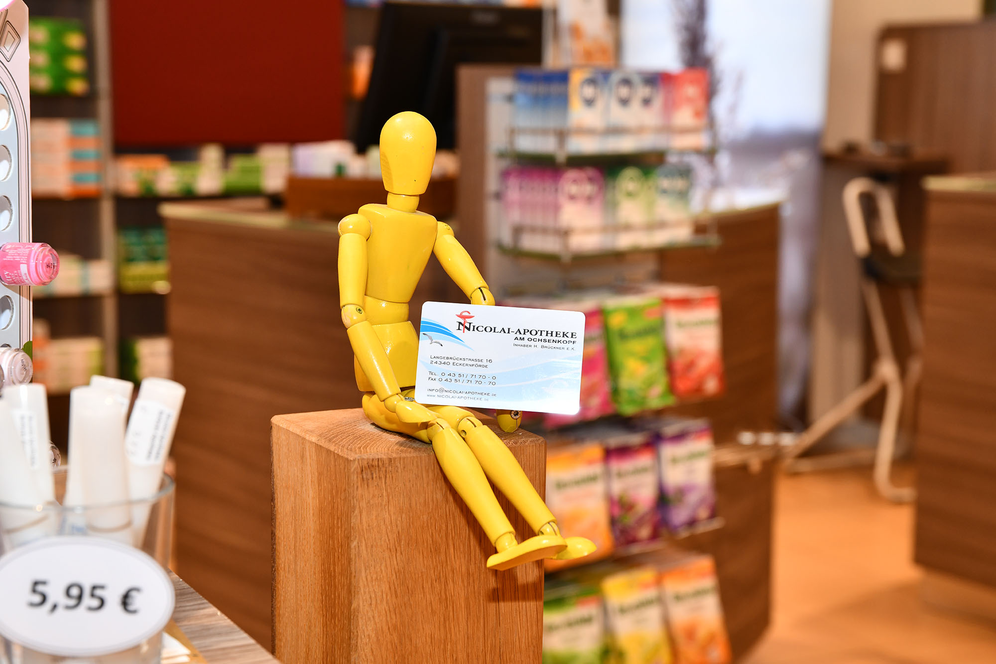 gelbes Holzmännchen mit der Kundenkarte. Im Hintergrund unscharf die Apotheke