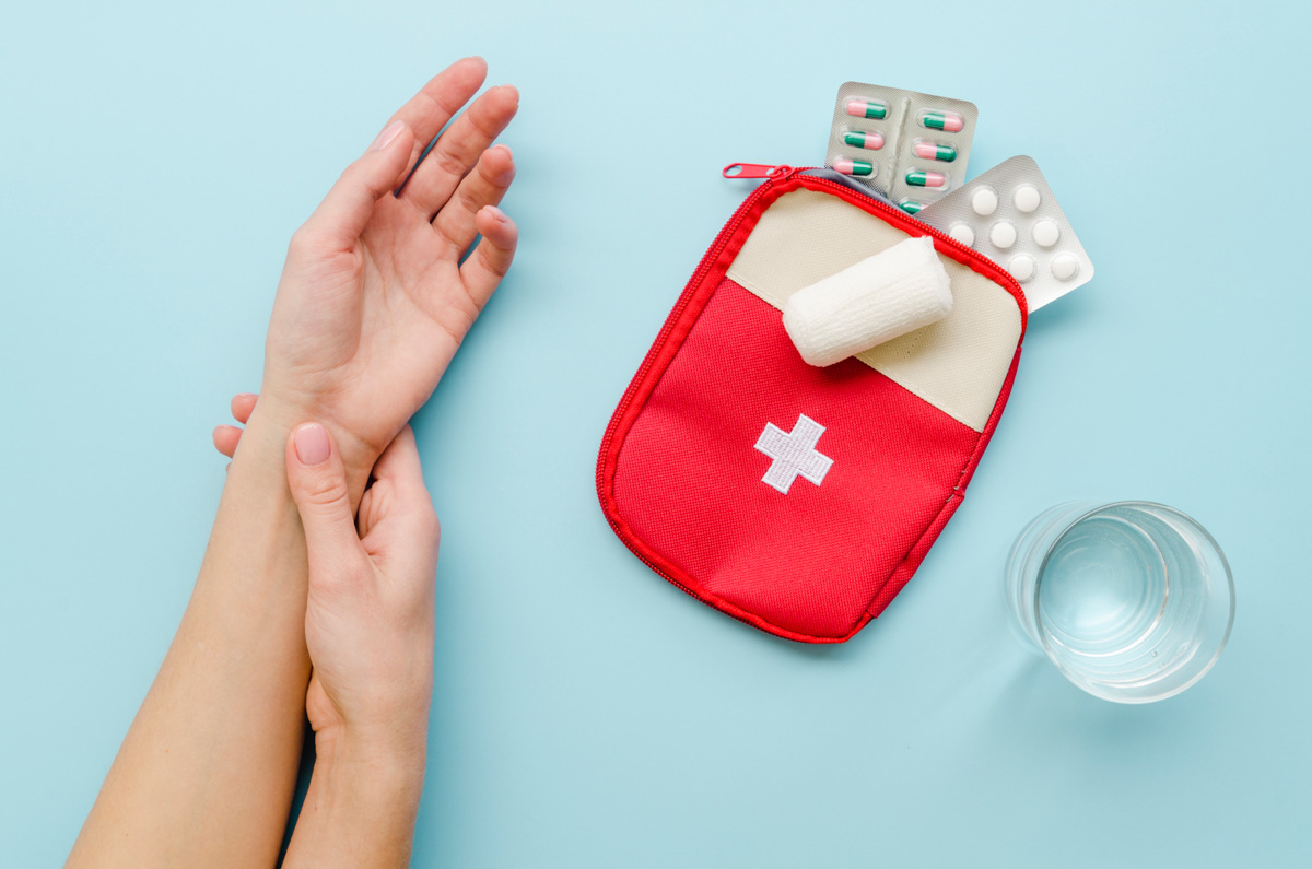 Handgelenk wird mit der anderen Hand abgetastet neben einem roten Erste-Hilfe-Set mit Verband und Medikamentenblistern sowie einem Glas Wasser